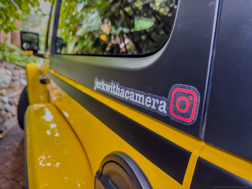 Close-up of a yellow Jeep Wrangler JL with a custom Instagram handle decal, surrounded by lush greenery.