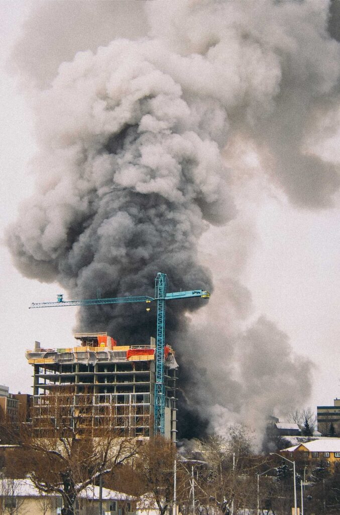 High-rise construction site with a tower crane in front of a large column of dark smoke rising into the sky.