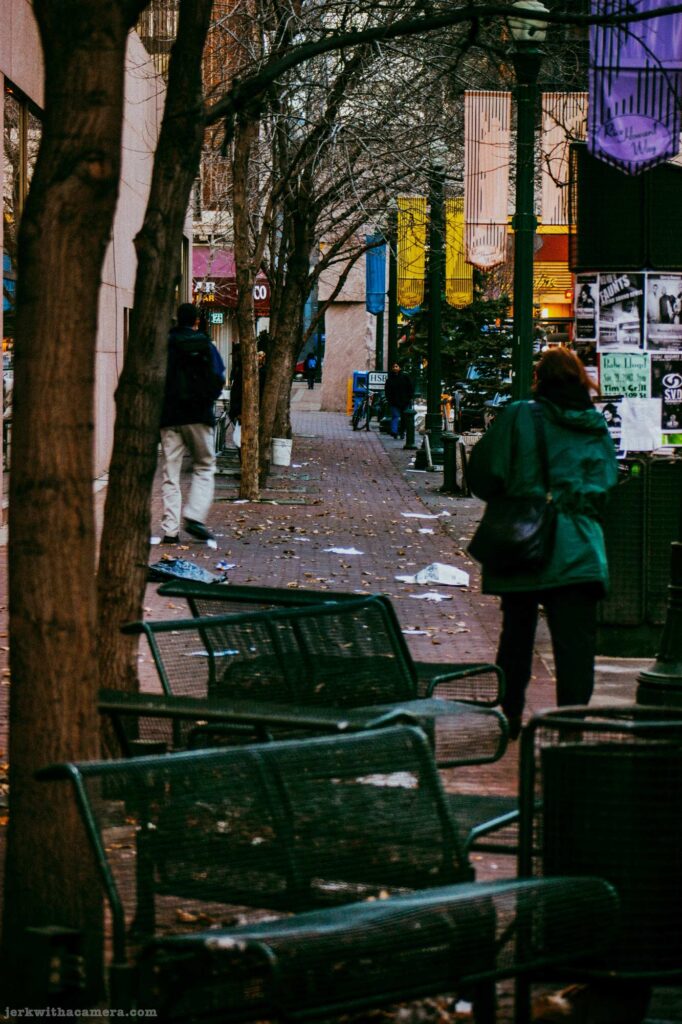 People walking along a downtown sidewalk lined with benches and trees