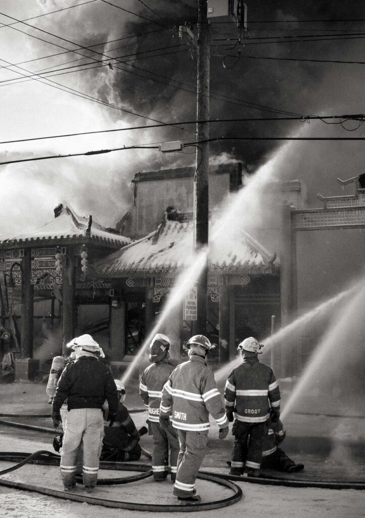 Firefighters stand near hose lines while water streams are directed at a building fire.