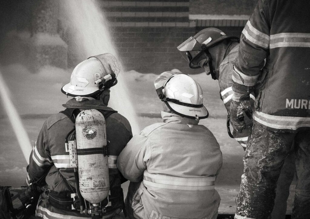 Two firefighters sit on snow-covered ground beside a hose line while another firefighter stands nearby.