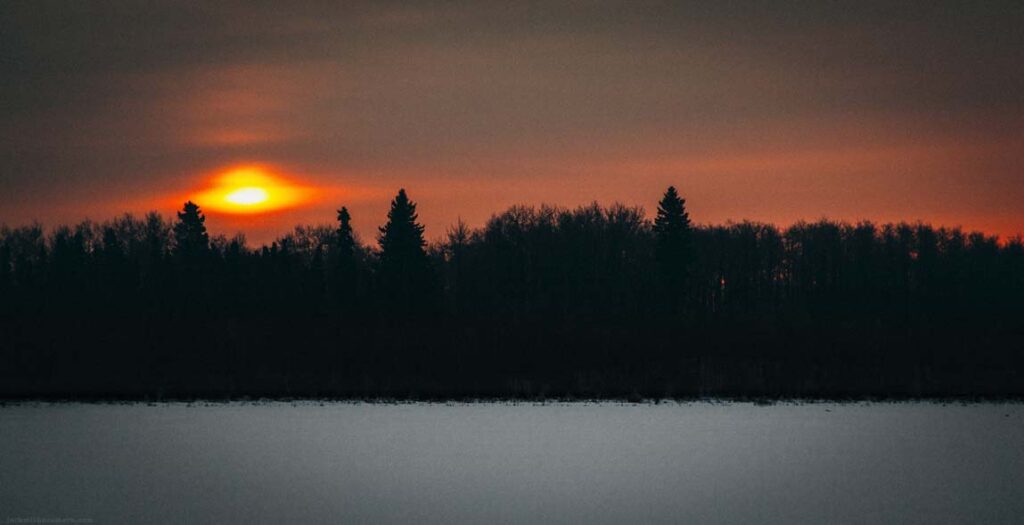 Sun setting behind a tree line across a frozen snow-covered lake