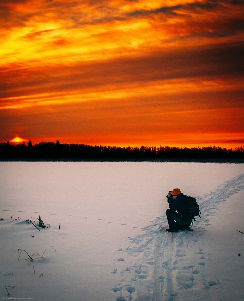 Person kneeling on a frozen lake photographing a sunset in winter