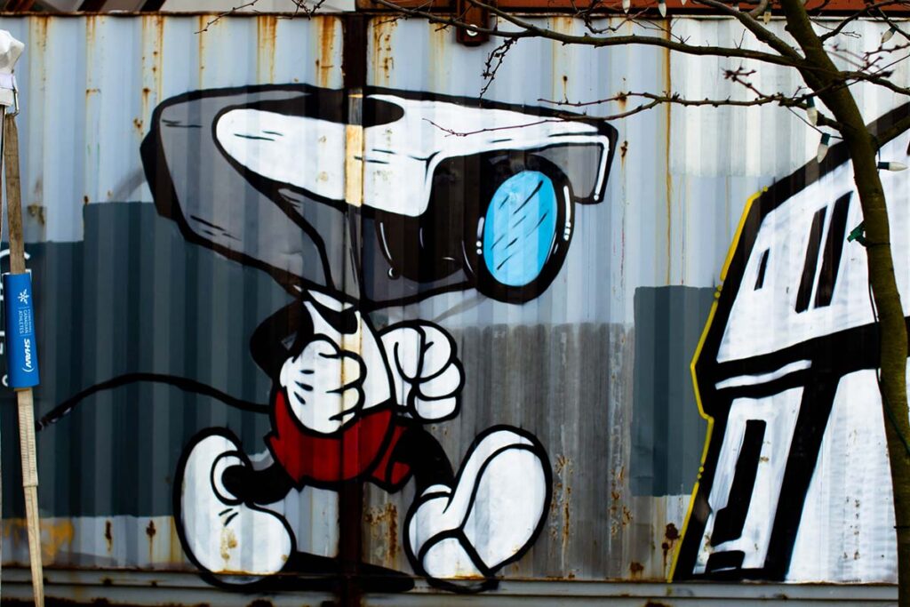 Graffiti of a cartoon character with a security camera for a head painted on a corrugated metal wall.