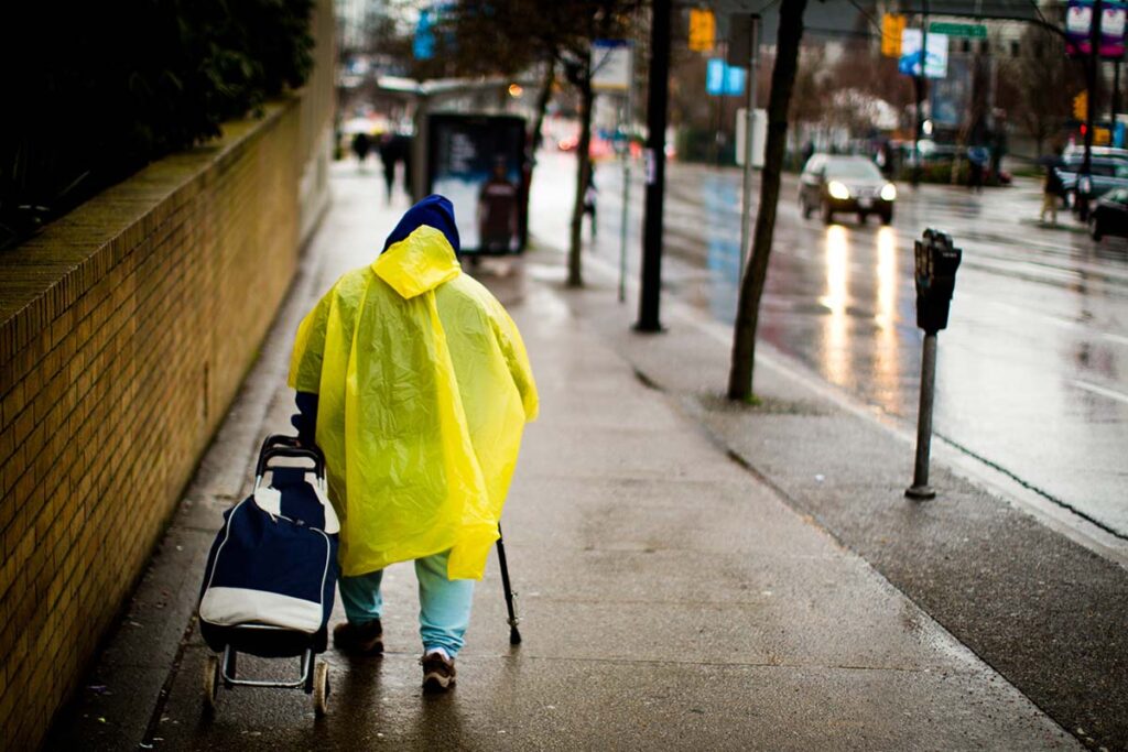 A person walking down a wet sidewalk in a bright yellow rain poncho, pushing a shopping trolley, on a rainy urban street.