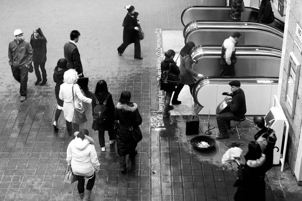 A busy urban scene with pedestrians passing a musician playing a hang drum near escalators.