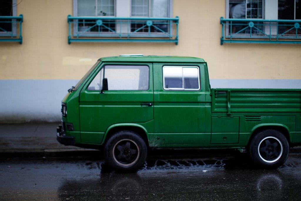 A vibrant green vintage Volkswagen pickup truck parked on a wet street with a pastel yellow building in the background.