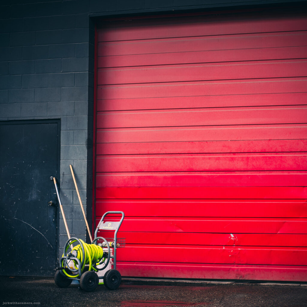 A bright red garage door with cleaning equipment leaning against a black wall.