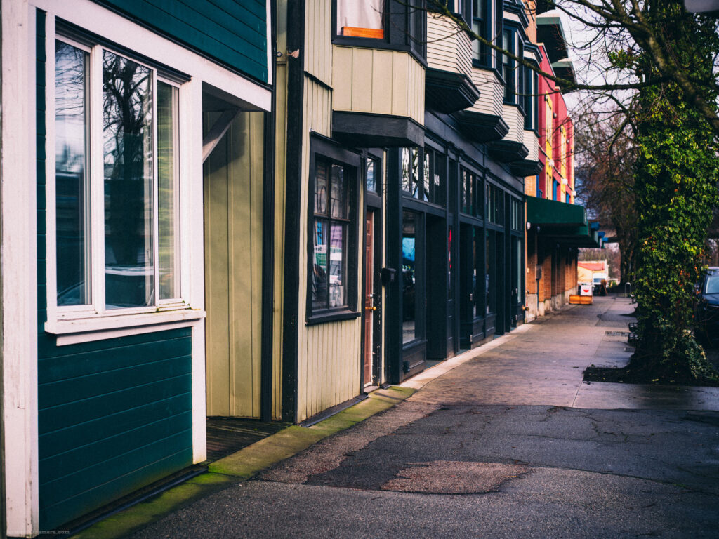 A row of colourful storefronts with a sidewalk framed by greenery.