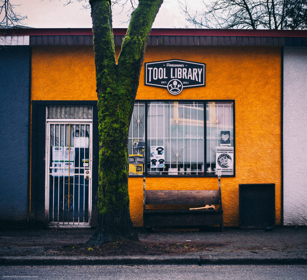 A bright yellow wall with the Vancouver Tool Library sign, framed by a moss-covered tree.