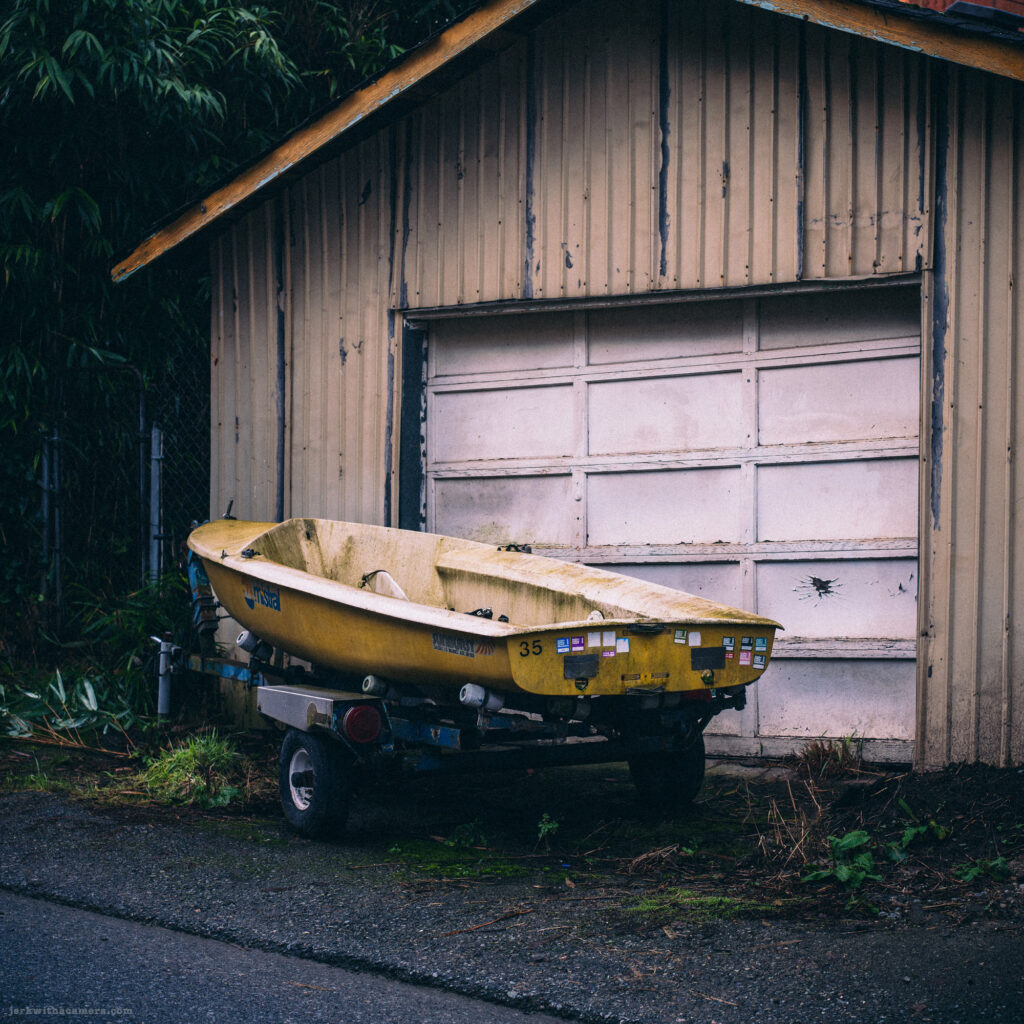 A small yellow boat on a trailer parked in front of a weathered garage.
