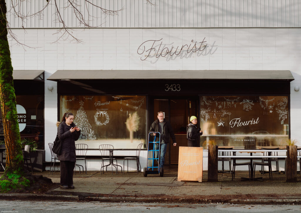 A storefront of the Flourist Mill & Bakery with people standing outside, including a delivery person and a woman checking her phone.