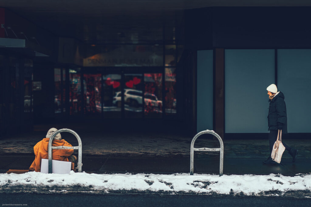 A person sitting on the sidewalk near a metal bike rack while another person walks by, with snow on the ground and a glass storefront in the background.