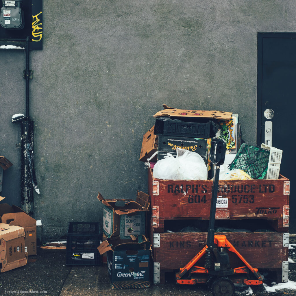 Stacked wooden crates filled with discarded boxes and plastic bags, with an orange pallet jack in front of a grey wall.