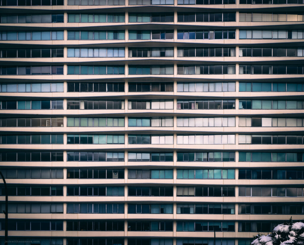 A multi-story building with rows of glass windows and balconies, some open and some with curtains.