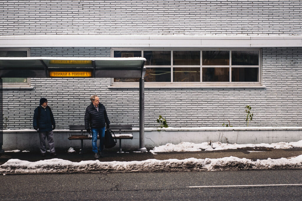 Two people waiting at a bus stop under a yellow sign, with a white brick wall and snow on the ground
