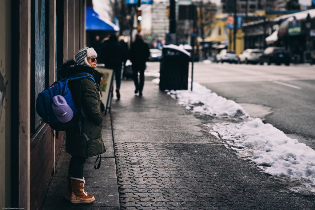 A person wearing winter gear and a large backpack stands on a sidewalk with snow, while pedestrians walk in the background.