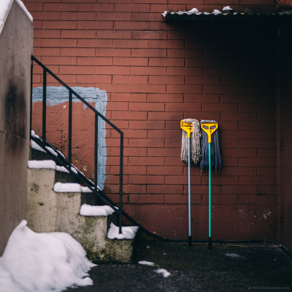 Two yellow mops leaning against a red brick wall next to a metal staircase with snow.