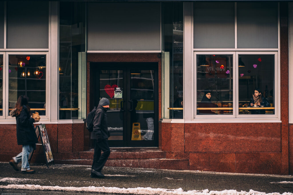 A café window with two people sitting inside, while pedestrians walk by outside on a snowy sidewalk.