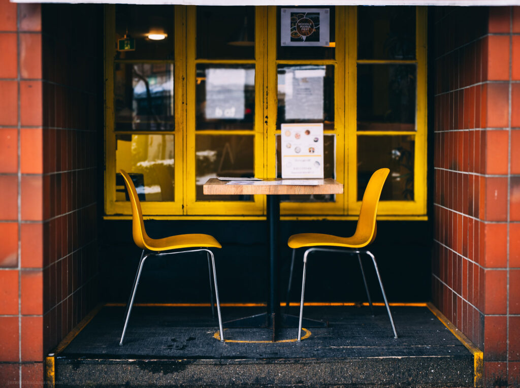 A small outdoor café area with two yellow chairs and a wooden table, set against a bright yellow window.