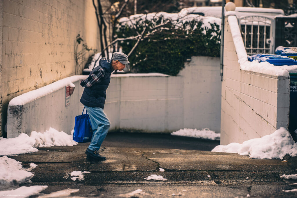 An elderly man wearing a plaid shirt and puffer vest walks uphill on a snow-lined driveway, carrying a blue shopping bag.