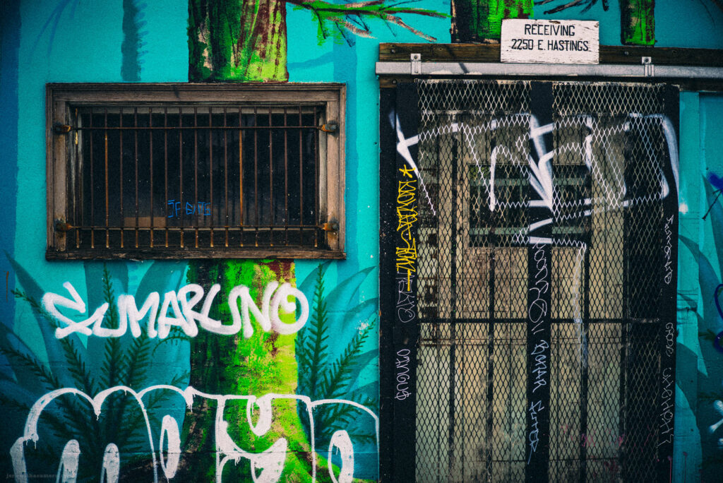 A graffiti-covered metal gate with a sign reading "Receiving 2250 E. Hastings," framed by a teal and green wall with barred windows.