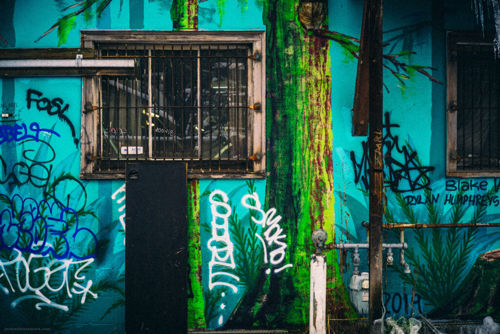 A barred window on a teal wall covered in graffiti, with pipes, cables, and a black panel partially obscuring the lower portion.