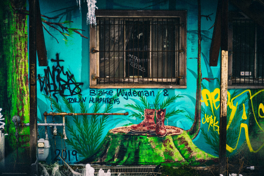 A graffiti-covered teal wall with a painted tree stump and a pair of red boots, alongside a barred window and utility pipes.