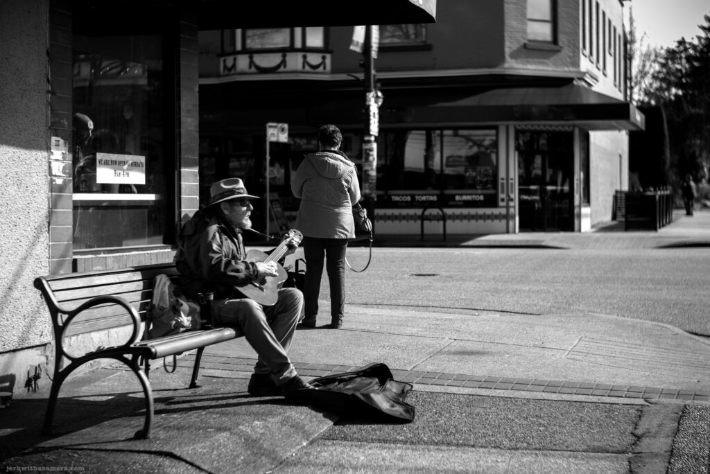 Man playing guitar on a bench while another person stands nearby at a street corner