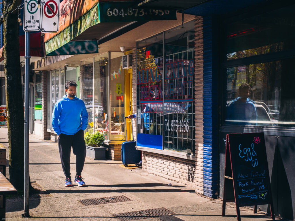 Man in a blue hoodie walking past storefronts with signs and reflections in the window.