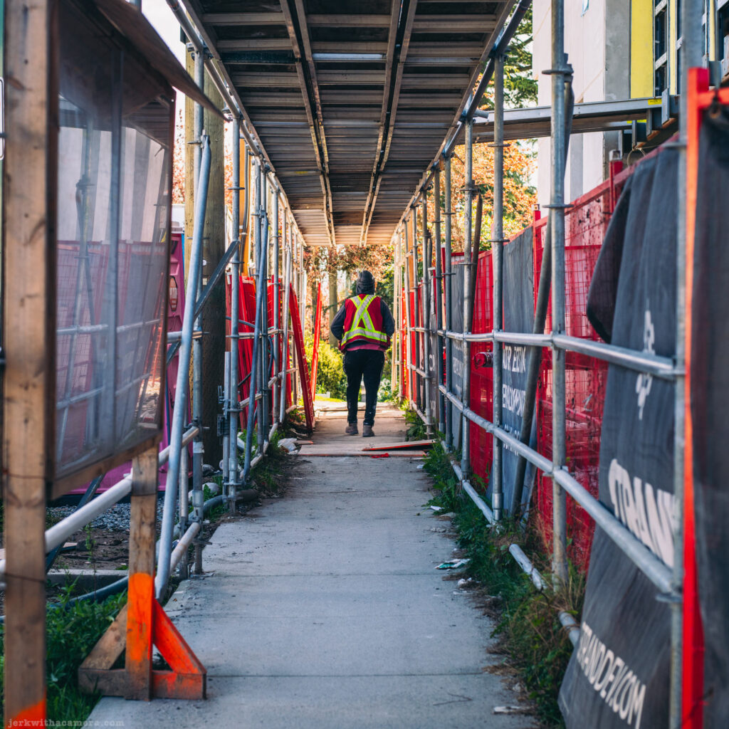 Construction worker walking under scaffolding on a narrow sidewalk path.