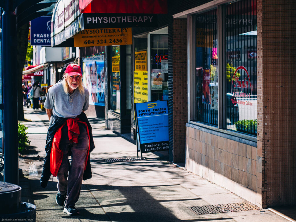 Older man in a red cap walking along a sidewalk past storefronts on Vancouvers Fraser Street