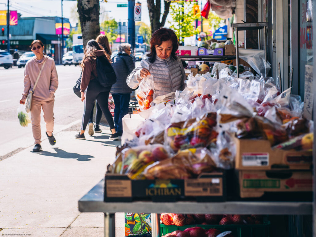 Woman selecting produce outside a Fraser Street grocery store while pedestrians walk past.