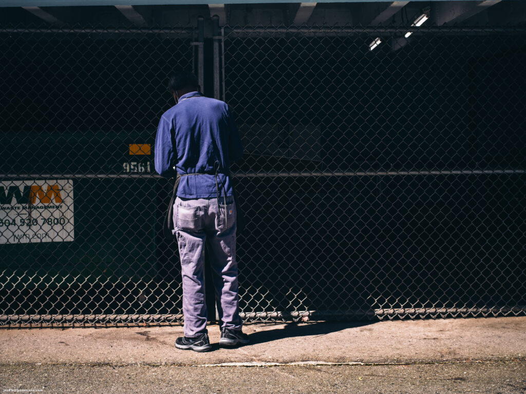 Person in blue overalls standing with their back to a chain link fence near a dumpster.
