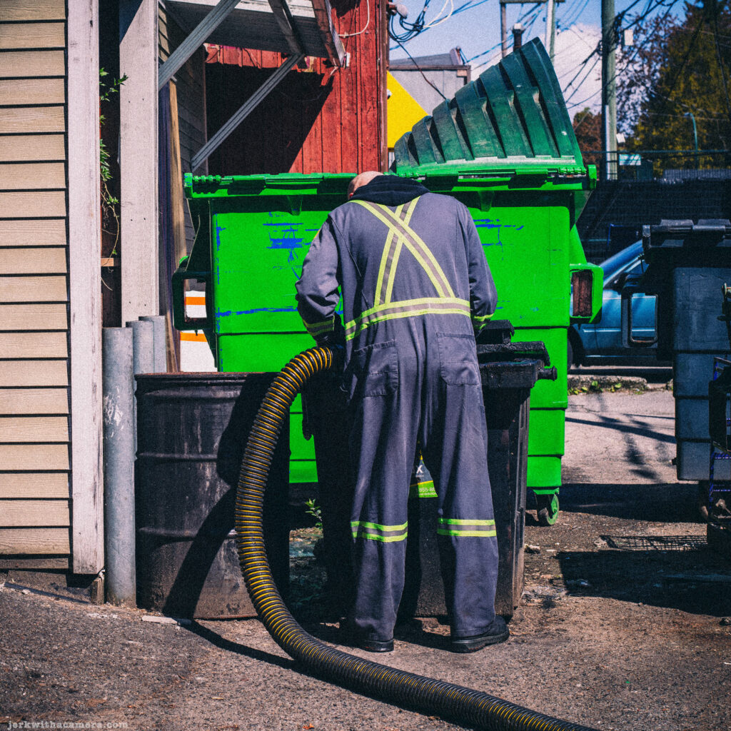 Worker in reflective coveralls cleaning or inspecting a green dumpster.