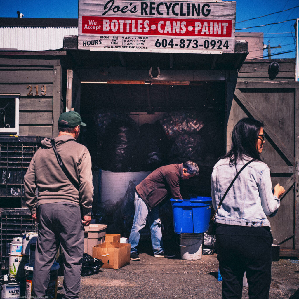 People standing outside a bottle depot labeled “Joe’s Recycling” with bins and bags visible.