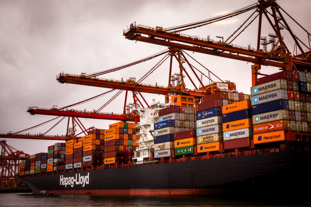 The cargo ship Hapag-Lloyd is docked at a shipping terminal with cranes and shipping containers stacked high.