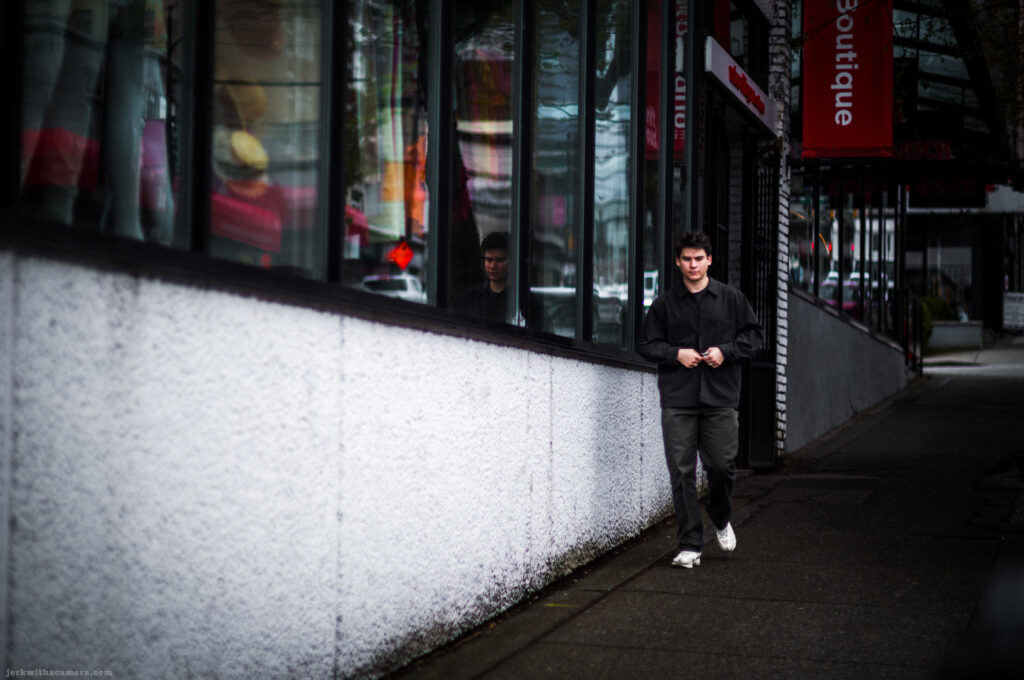 Person walking on a city sidewalk in front of a white stucco wall with reflective windows.