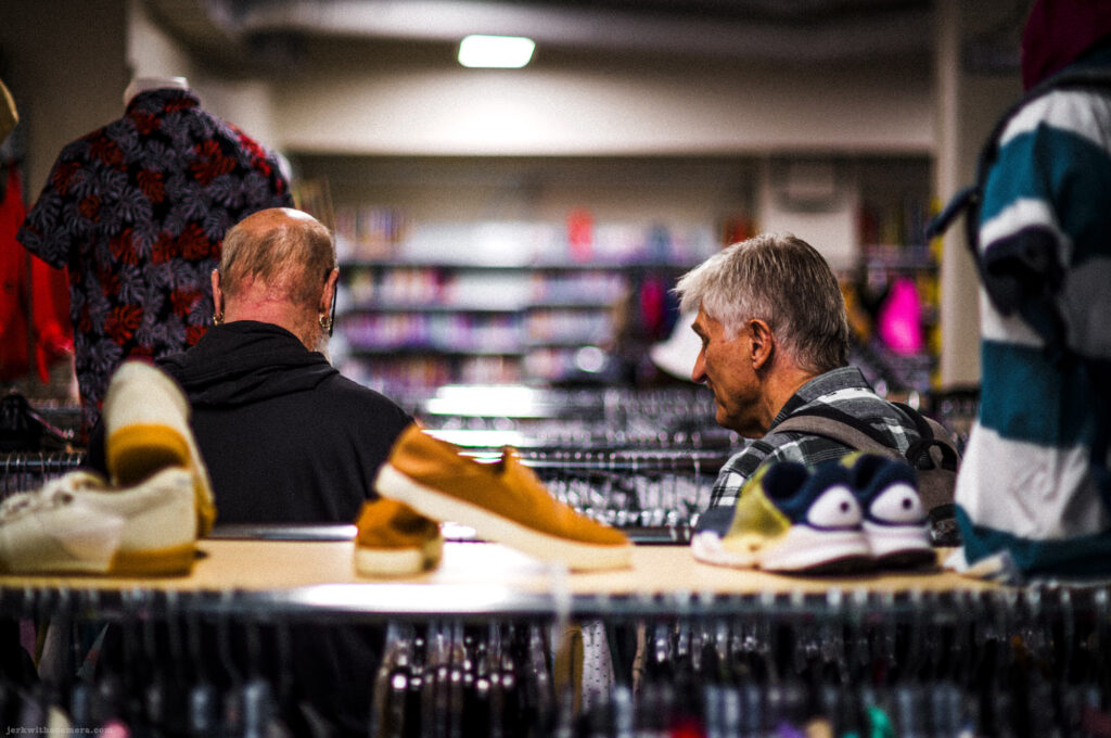 Two men sitting and talking in a secondhand clothing store surrounded by racks of clothes and shoes.