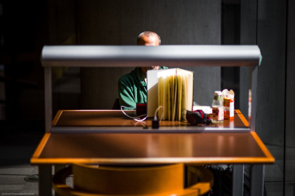Person seated at a library desk partially obscured by a desk lamp and open book.