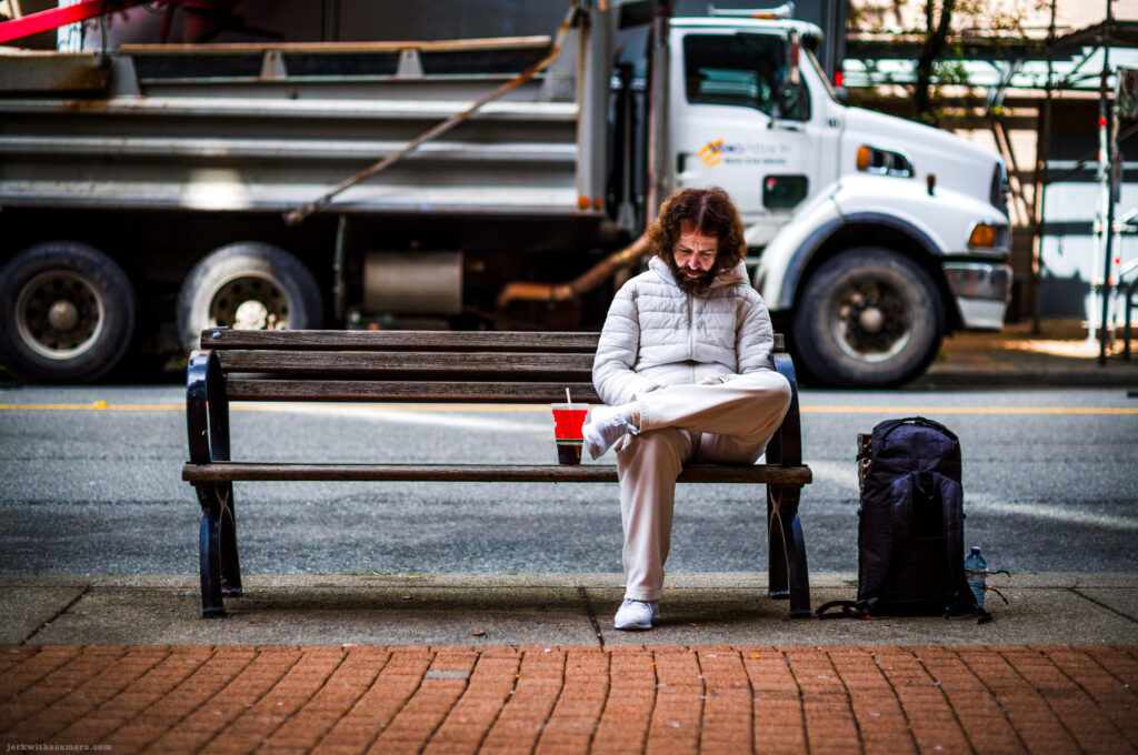 Man sitting on a bench with a drink cup and a backpack next to a large truck.