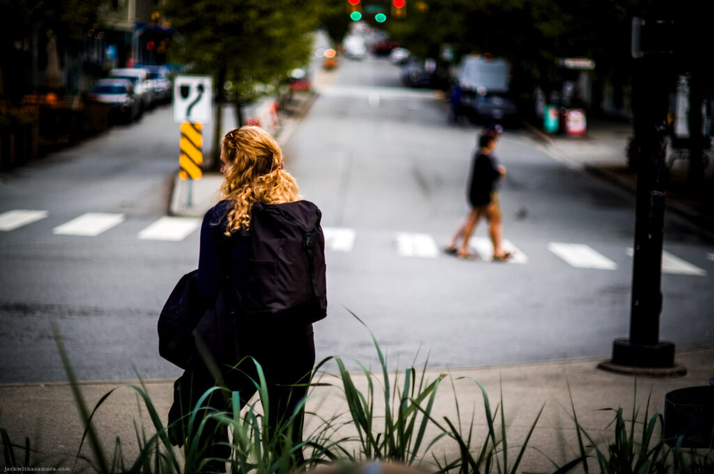 Woman with a backpack looking at a city street while two others cross in the background.