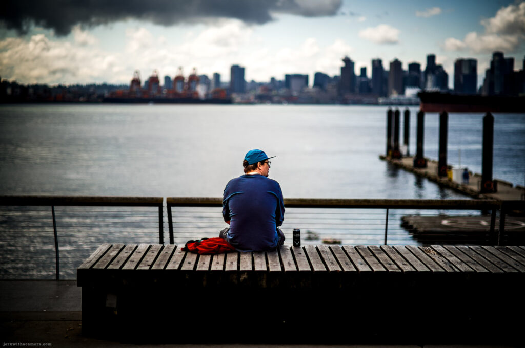 Person in blue clothing sitting on a wooden bench facing the Vancouver skyline across the water.