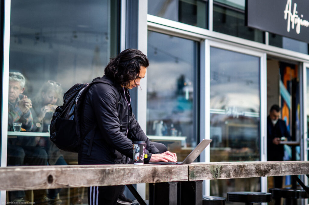 Man with a backpack typing on a laptop at a counter outside a cafe, with people visible through the window behind him.