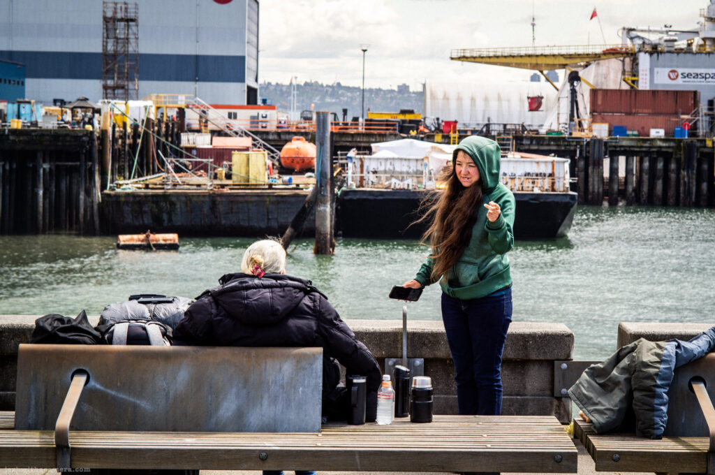 Two people by the water near an industrial dock, one sitting and one standing with a hood up.