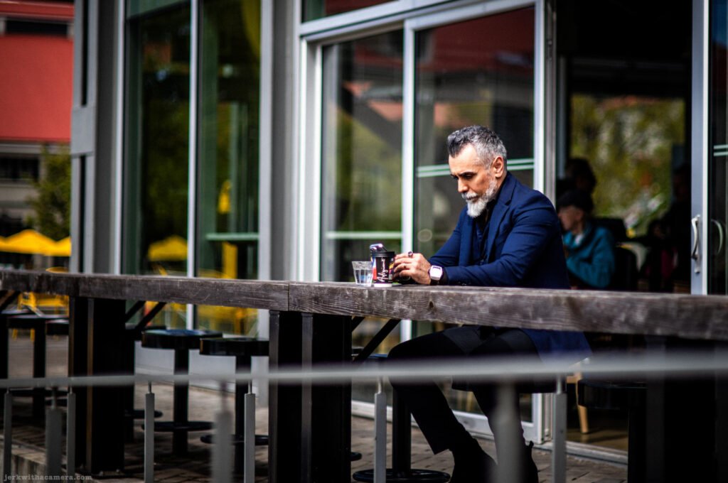 Man with white beard sitting alone at a wooden counter outside a cafe, holding a phone and coffee cup.