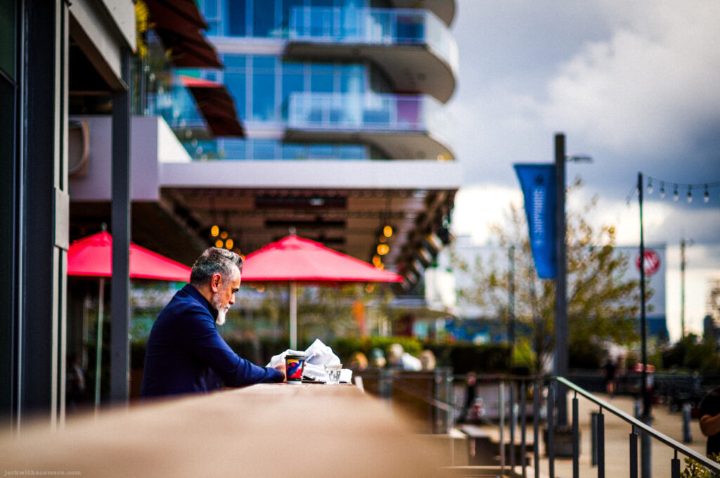 Man with white beard sitting alone at a wooden counter outside a cafe, holding a phone and coffee cup.