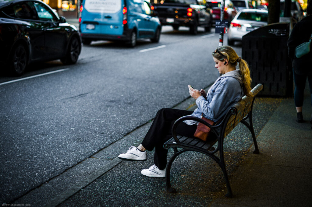 Woman sitting on a sidewalk bench looking at her phone with traffic passing nearby.