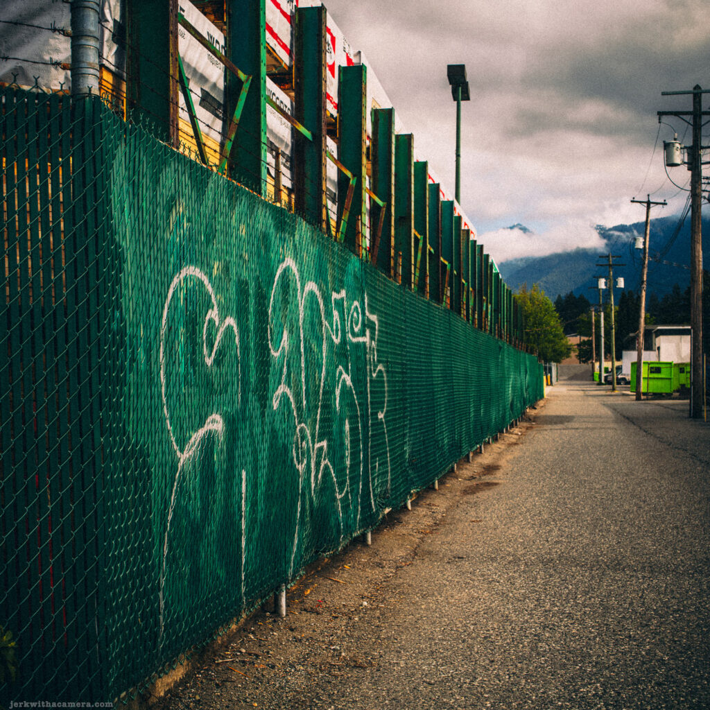 Green fence with graffiti beside a road
