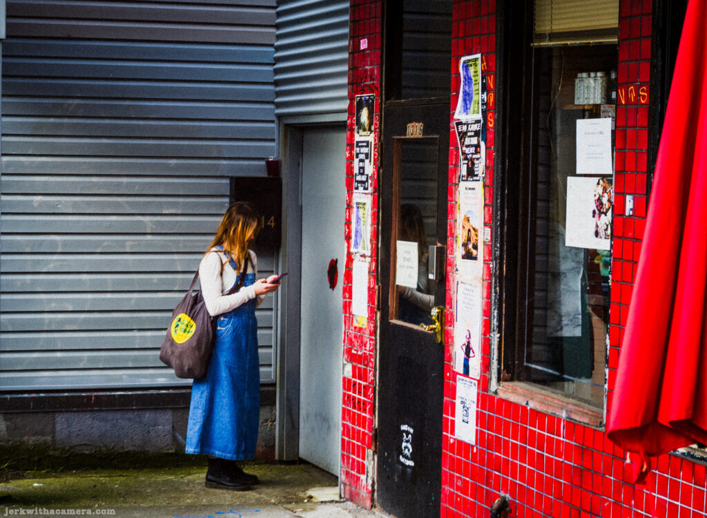 A woman stands outside a red-tiled building while looking at her phone.